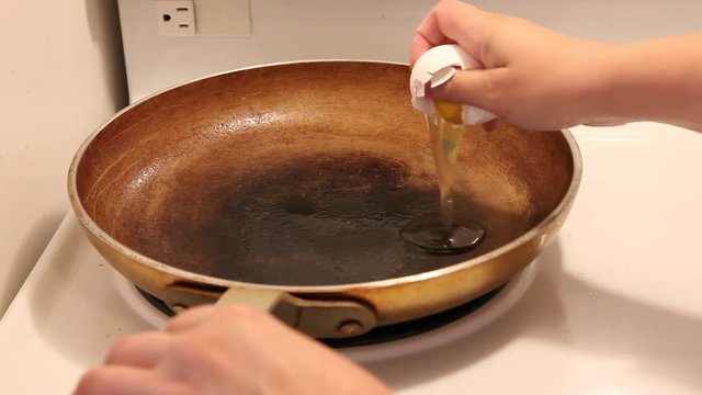 A hand tries to crack an egg into a skillet with one hand but gets shell in the pan. 