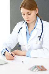 Doctor woman filling up prescription or medical record form while  sitting near window in clinic or hospital. Medicine and healthcare concept. Physician at work