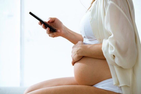 Pregnant woman texting with her smartphone sitting at home.