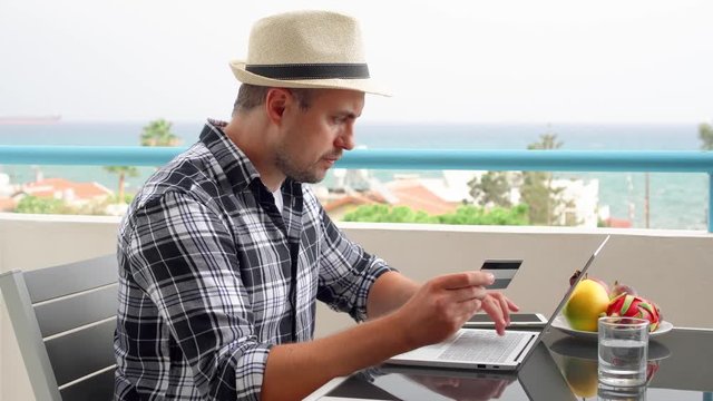 Businessman sit on balcony use laptop at tropical beach resort. Young professional male freelancer shopping online with credit card on computer from remote home office. Sea and palm trees on