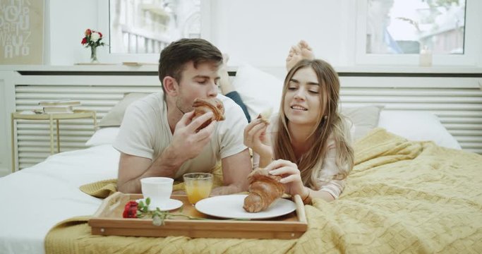 In the morning a cute couple have breakfast in bed eating French croissants and have a conversation , very cozy atmosphere.