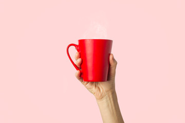 Female hand holding a red cup with hot coffee or tea on a light pink background. Breakfast concept...