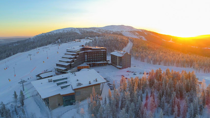 AERIAL: Flying around hilltop skiing resort surrounded by ski slopes in winter