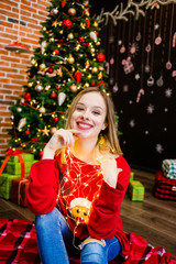 A comely girl in a red sweater sitting at a tree and holding garlands in her hands.