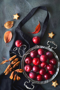 Vintage Metal Plate With Red Plums And Scattered Red And Orange Autumn Leaves On Black Apron