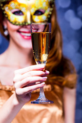 Style redhead girl in golden dress and mask keep glass of drink on gray background with bokeh