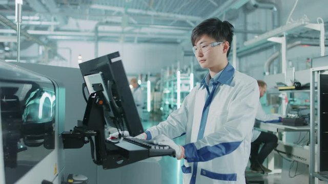 On High Tech Factory Asian Engineer Uses Computer for Programming Pick and Place Surface Mount Electronic Machinery for Printed Circuit Board Assembly Line. 
