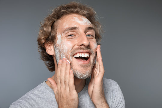 Young man washing face with soap on grey background