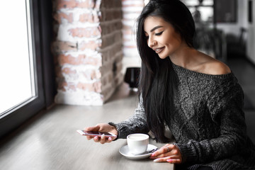 Beautiful girl uses a phone and drinks coffee, sitting in a cozy cafe.