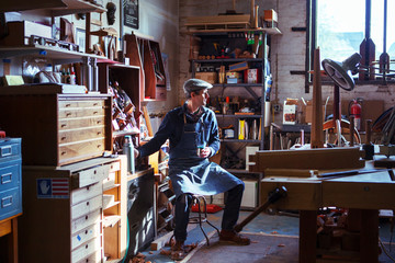 Fototapeta premium Carpenter holding drink looking away while sitting on stool in workshop