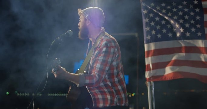 Medium shot of male singer in a plaid shirt singing and playing guitar on a urban rooftop at night with the US flag in the background.
