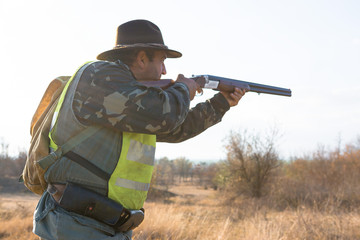 A hunter in a hat with a gun in camouflage and a reflective vest in the steppe