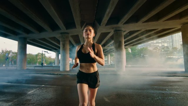 Portrait Shot of a Beautiful Fitness Girl in Black Athletic Top and Shorts Jogging Through a Smoky Street. She is Running in an Urban Environment Under a Bridge with Cars in the Background.