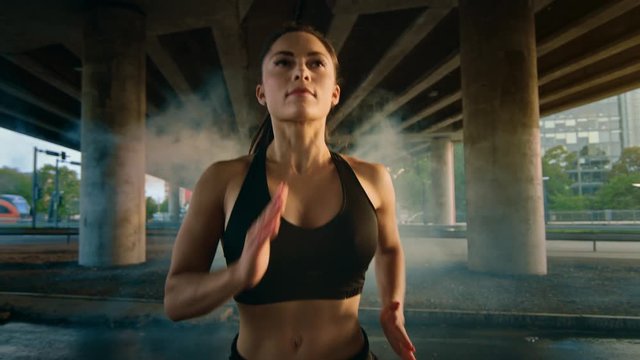 Close Up Portrait Shot of a Beautiful Confident Fitness Girl in Black Athletic Top and Shorts Jogging Through a Foggy Street. She is Running in an Urban Environment Under a Bridge with Cars