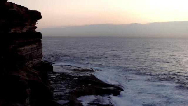Wide shot of two boats passing at sunrise with waves crashing against cliffs