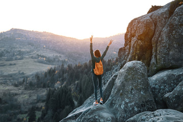 woman hiker with backpack stands on edge of cliff against background of sunrise. Woman is meditation and greeting a sun