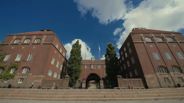 People walk in front of the entrance to KTH Royal Institute of Technology. Large brick buildings, an iron fence and tall flag poles frame the entryway of the campus. Low, wide angle view.