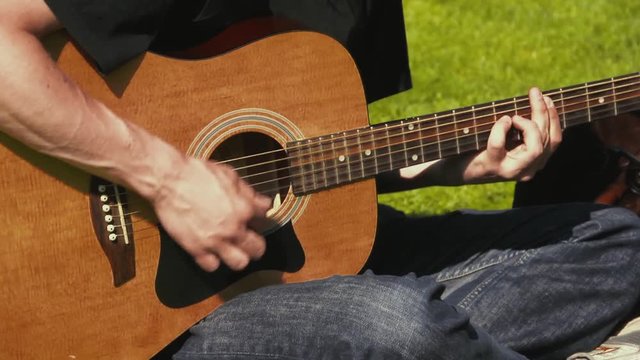 Close up of a young man playing acoustic guitar while sitting outside on the grass. Man wearing jeans and black shirt strumming a guitar in the sunshine.