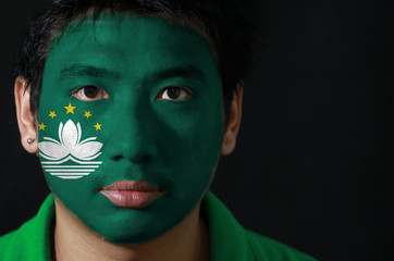 Portrait of a man with the flag of the Macau painted on his face on black background, green with a lotus and stylised Governor Nobre de Carvalho Bridge and water with star.