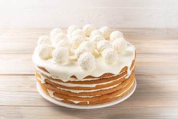 The festive biscuit cake made of white cream is decorated with round candies with coconut chips on the table on a light background. Copy space. Close-up