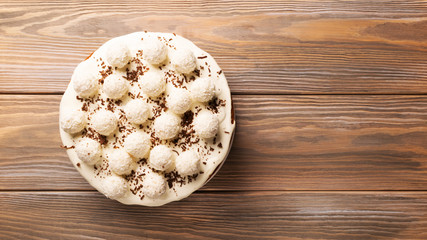 Biscuit cake poured with white cream is decorated with round candies with coconut chips on a wooden background. Copy space. View from above. Flat layout