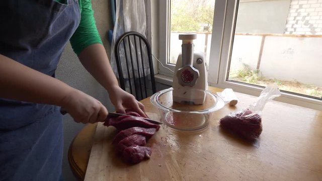 Female cook in apron is cooking sweet peppers stuffed with meat in kitchen. girl cuts the beef into large pieces, puts it in meat grinder. Pushes pieces into meat grinder, mixes minced meat.