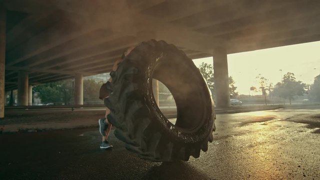 Beautiful Confident Fitness Girl in Black Athletic Top is Doing Exercises in the Street. She's Flipping a Big Heavy Tire in an Urban Environment.
