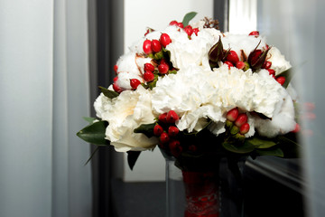 Wedding bride's bouquet of white flowers, red berries and greenery standing in a vase on the windowsill in soft-focus in the background. wedding attributes near the window. bridal bouquet.