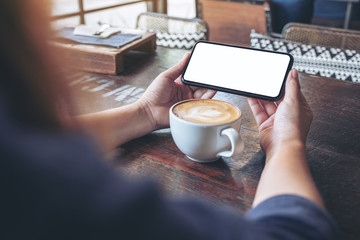 Mockup image of a woman holding and using a black mobile phone with blank screen horizontally for watching with coffee cup on wooden table