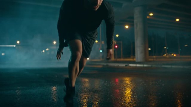 Strong Muscular Fit Young Man Starts Sprinting on a Rainy Evening. He is Running in an Urban Environment Under a Bridge with Cars in the Background.