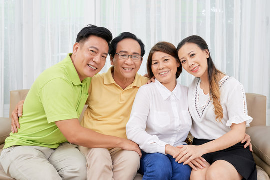 Hugging happy Vietnamese family sitting on comfortable sofa at home and smiling at camera