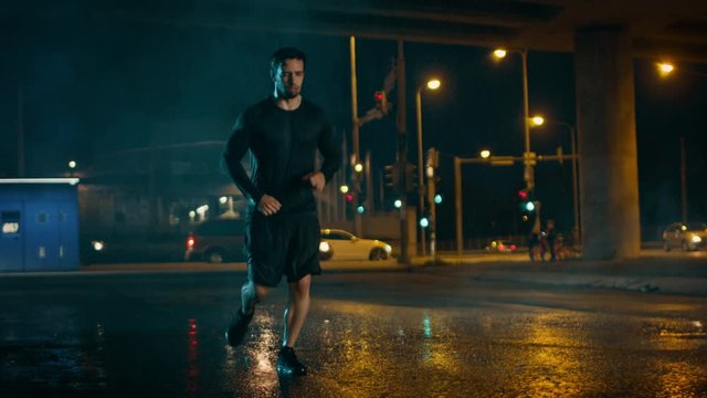 Athletic Muscular Young Man in Sports Outfit is Jogging in the Street on a Rainy Evening. He is Running in an Urban Environment Under a Brindge.