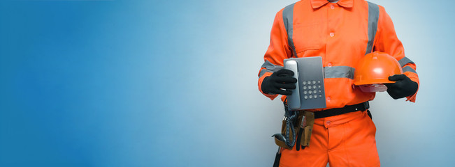 Builder worker is holding in hand a handset phone and hard hat isolated on blue background with copy space. Call to a repair man concept. Contact us.