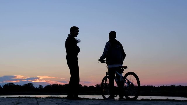 Silhouette of a man and a teenager with a bicycle being photographed against the background of the morning sky on the bank of the river or lake. People take a selfie on the phone at dawn. Life style.