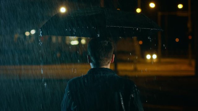 Backshot of a Young Caucasian Shorthaired Man, Wearing a Jeans Coat Walking in the Rain Under an Umbrella. He's Walking at Night in the City.