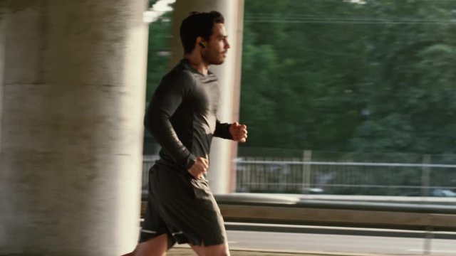 Athletic Young Man in Earphones and Sports Outfit is Jogging in the Street. He is Running in an Urban Environment Under a Brindge with Cars in the Background.