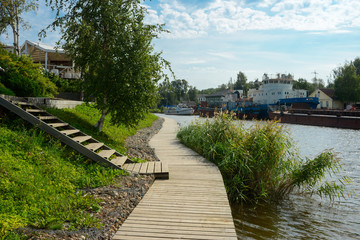 Fototapeta premium Rustic wooden walkway along the river. Countryside. Old rusty ships in the background. Summer time