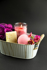 Wellness spa therapy pampering products on black background, pink candle burning, cozy mood. Pink bath bomb, hand made soap, pink sponge, flowers for decoration in white metal vintage basket.