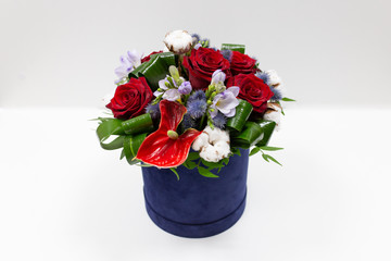 A bright bouquet of red flowers (rose and anthurium) with green leaves, Alstroemeria and cotton flowers in a blue box for hats on a light background