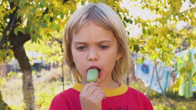 Excited elementary age girl eating tasty green fruit popsicle in summer. Happy cute child enjoying taste of fresh fruit ice cream outdoors on hot summer day, beautiful background.
