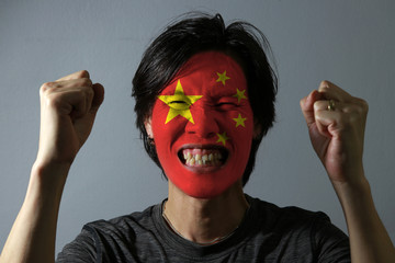 Cheerful portrait of a man with the flag of the China painted on his face on grey background. The...