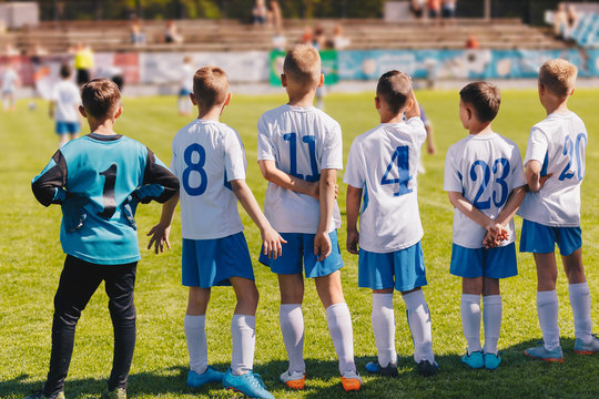 Children Sport Team Photo. Group of Young Boys Playing Soccer Tournament Match. Football Soccer Game For Children. Kids Soccer Players Standing Back on Bench and Watching Tournament Game