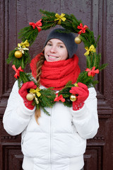 girl in red gloves and white jacket holding Christmas wreath in front of the door