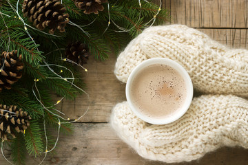 Women's hands in mittens hug a cup of cocoa on the background of fir branches with pine cones and a garland.