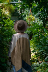 Fashionable young woman wearing in hat and poncho among tropical plants. Rear view.