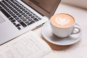 Laptop with a graph, schedule, a glass of coffee with heart ornament and copybook with notes, place for business work and productivity