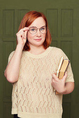 Indoor vertical shot of stupefied European red headed woman keeps hand on rim of spectacles, has unexpected gaze at camera, receives strange news, holds books, posing on classroom door background.
