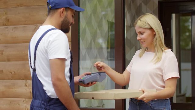 PAN shot of cheerful female customer receiving pizza from delivery man in uniform and paying with credit card