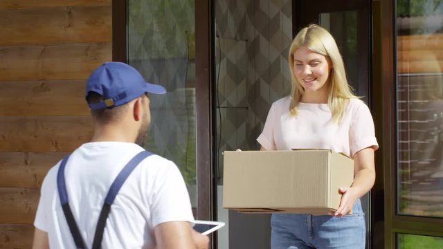 Medium shot of blond woman giving cardboard box for shipping to male courier and signing electronic invoice on tablet