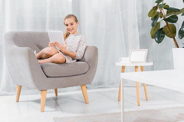 cheerful little child in earphones sitting on armchair with digital tablet at home